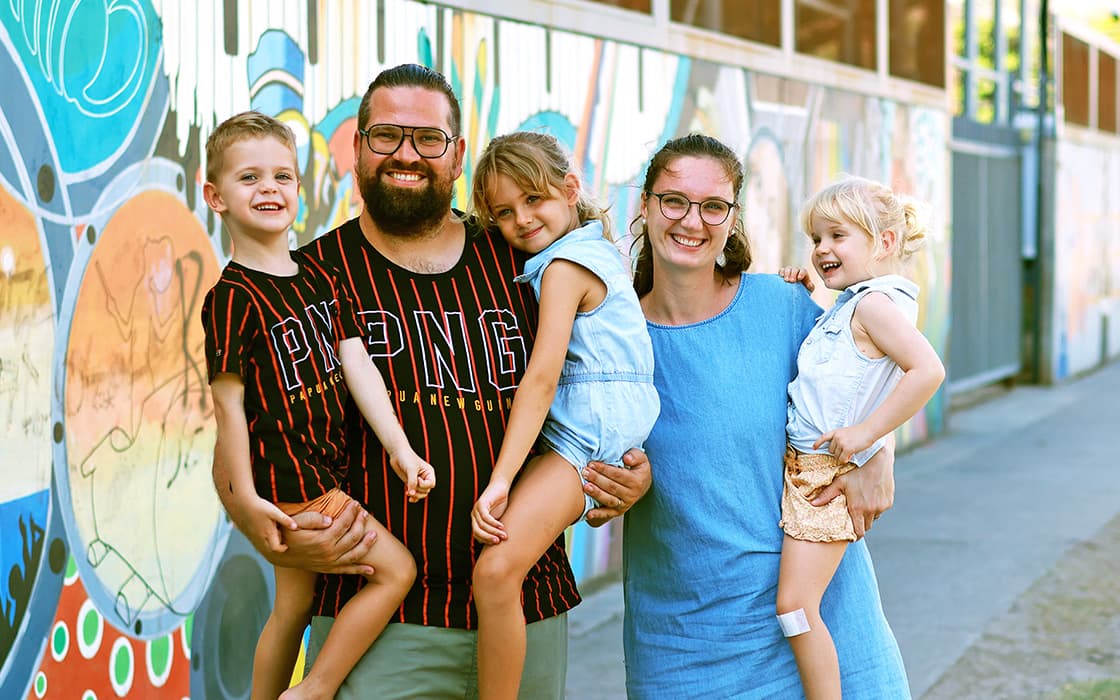 Eine lächelnde Familie mit drei kleinen Kindern, Sebastian Proß und Katharina Proß, posiert an einem sonnigen Tag vor einem bunten Wandgemälde.