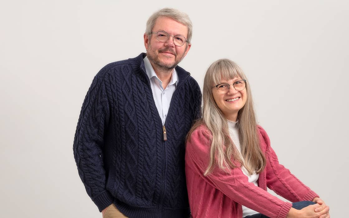 Der lächelnde Peter Rapp in einem dunklen Pullover steht neben der sitzenden Sigrun Rapp in einer rosa Strickjacke vor einem schlichten Hintergrund.