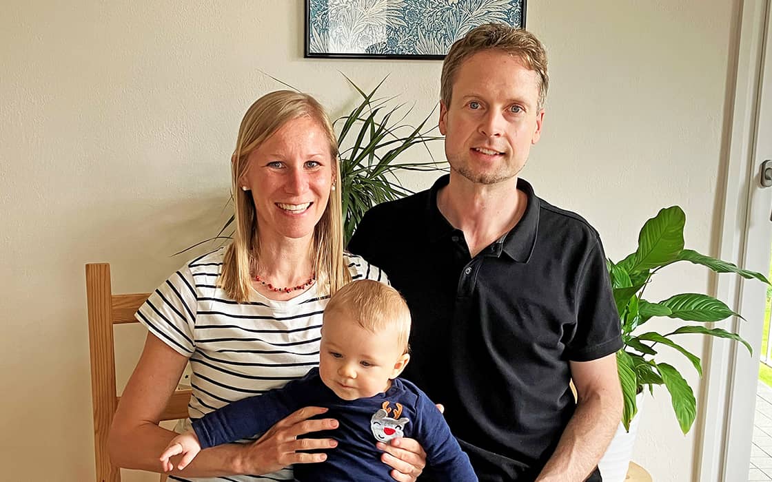 Ramona Kröger und Robert Kröger sitzen lächelnd zusammen, mit einem Baby auf dem Schoß, in einem Innenraum mit Grünpflanzen im Hintergrund.