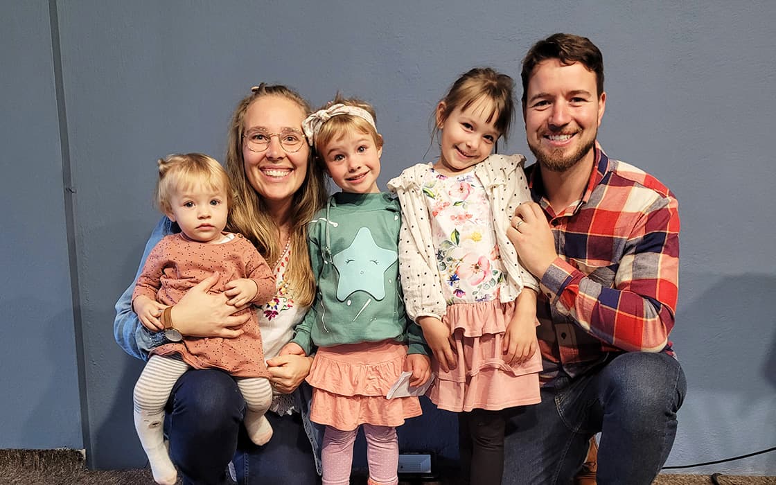 Eine lächelnde fünfköpfige Familie, zu der auch Tabea Ruf gehört, posiert zusammen mit ihren drei kleinen Töchtern vor einer blauen Wand.