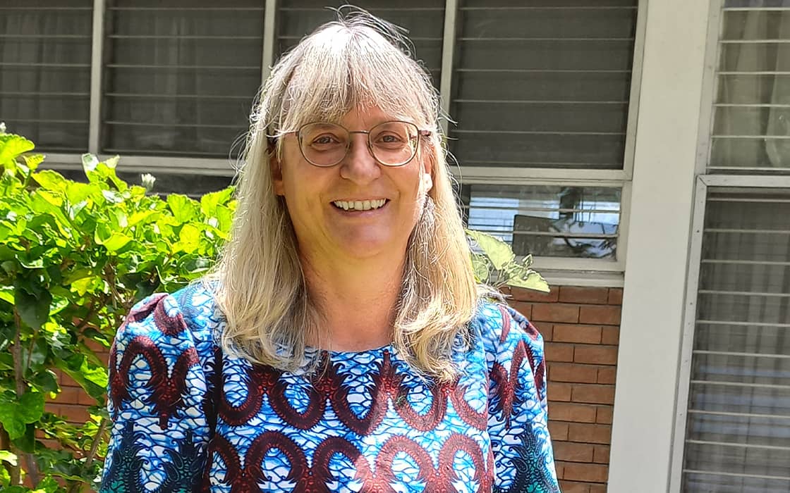 Margit Schwemmle, lächelnd, mit langen grauen Haaren und Brille, steht draußen zwischen grünen Büschen in der Nähe eines Gebäudes mit Fenstern.