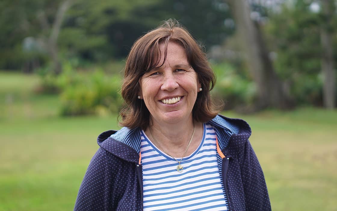 Brigitte Stamm, eine Frau mit braunem Haar, lächelt im Freien in einem gestreiften Hemd und einer marineblauen Jacke vor einem grünen Hintergrund.