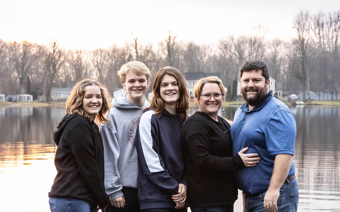 Anne-Christine Strauß und Samuel Strauß lächeln mit Freunden an einem ruhigen See bei Sonnenuntergang, Bäume und Häuser im Hintergrund.
