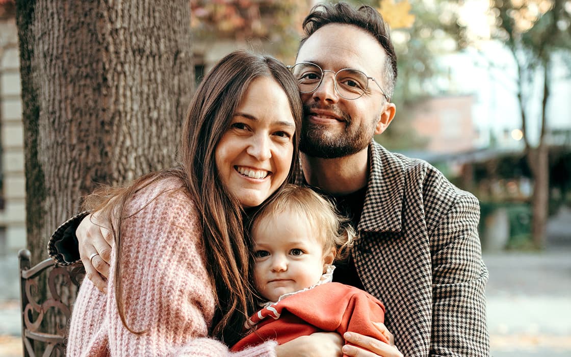 Benjamin und Julia Stute umarmen ein kleines Kind im Freien, ganz in warmer Kleidung, mit Bäumen und Blättern im Hintergrund.