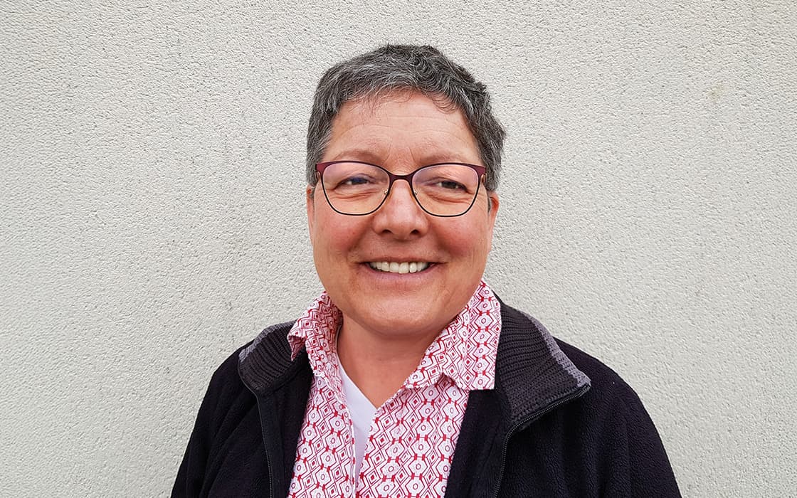 Evelyn Theurer, mit kurzen grauen Haaren und Brille, lächelt in einem gemusterten Hemd und einer dunklen Jacke gegen eine helle Wand.