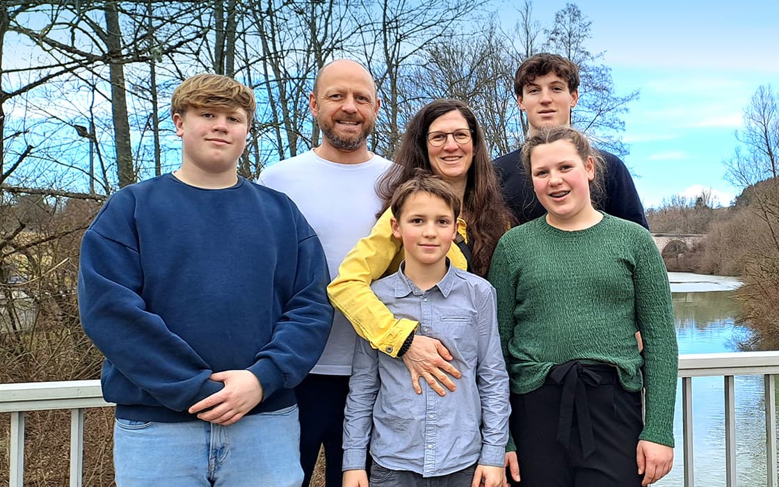 Susanne Trick und Stephan Trick lächeln mit ihrer sechsköpfigen Familie auf einer Holzbrücke, Bäume und Fluss im Hintergrund.