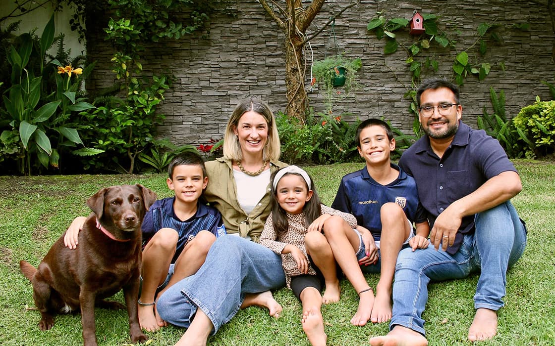 Eine fünfköpfige Familie und ihr brauner Hund sitzen lächelnd im Gartengras; Andrés Vergara strahlt zwischen Grünpflanzen und einer Steinmauer.