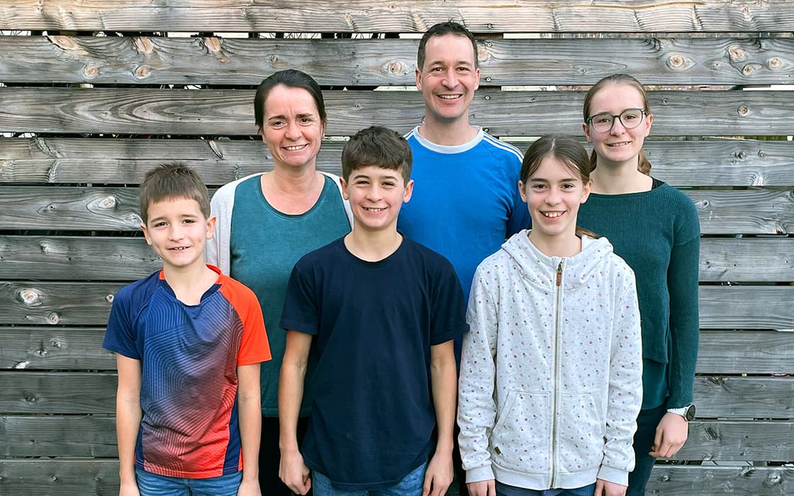 Sarah Zinser und Tobias Zinser lächeln mit ihren vier Kindern vor einem Holzzaun, drei Jungen und ein Mädchen vor ihnen.