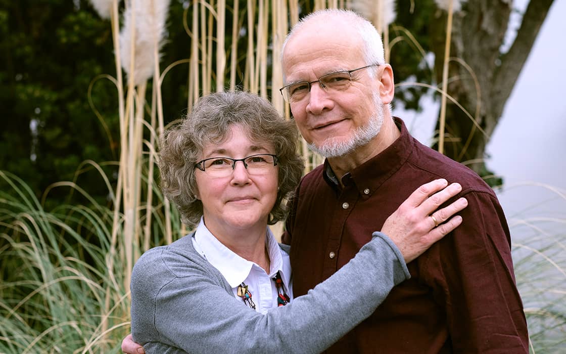 Elisabeth Bredow und René Bredow stehen dicht beieinander im Freien und lächeln inmitten von hohem Gras und Bäumen.