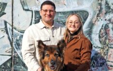 Anna Marasco und ein Mann lächeln im Freien mit einem großen Hund vor einer bunten, mit Graffiti beschmierten Wand.