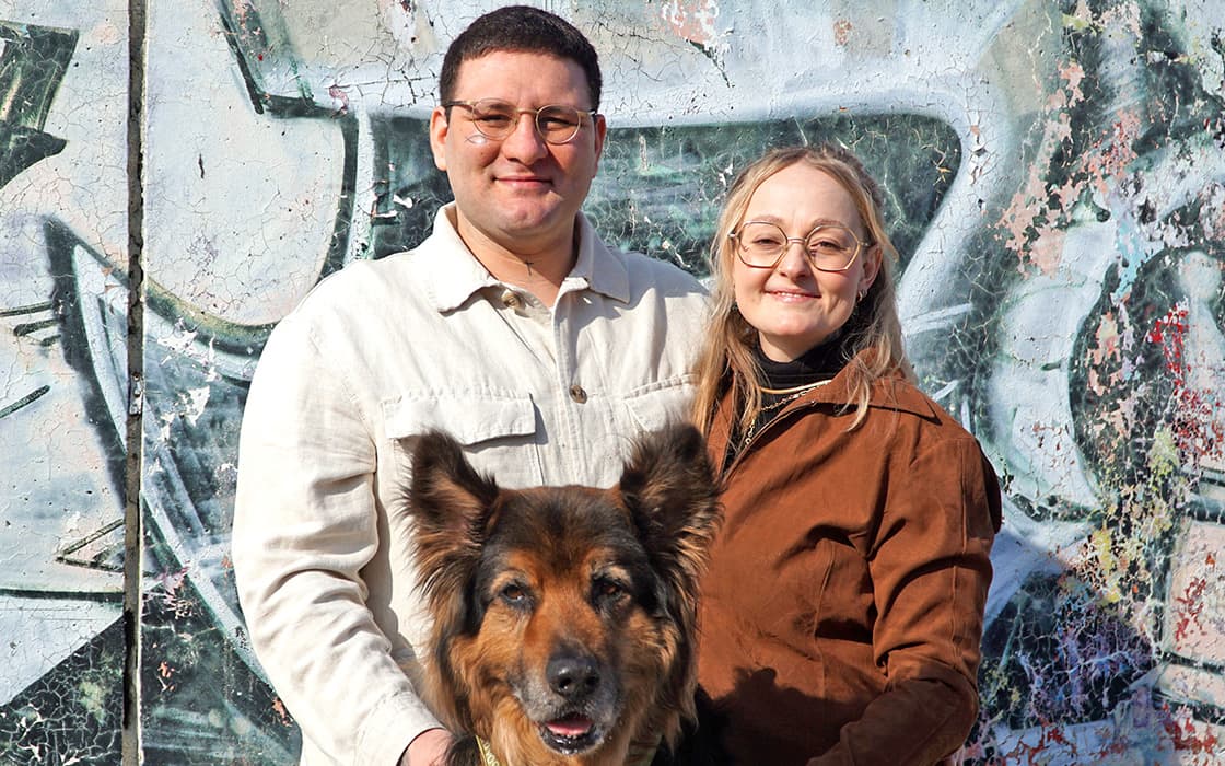 Anna Marasco und ein Mann lächeln im Freien mit einem großen Hund vor einer bunten, mit Graffiti beschmierten Wand.