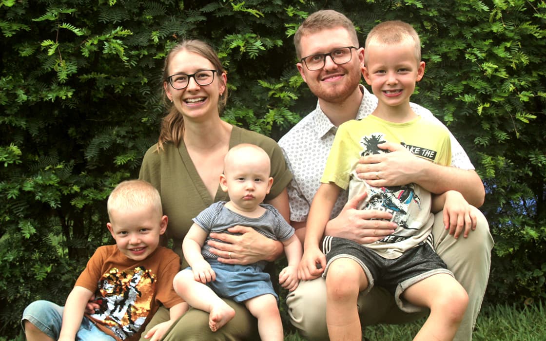 Eine lächelnde fünfköpfige Familie, darunter Esther Stoll und Manuel Stoll, posiert im Freien vor grünen Büschen mit ihren drei kleinen Kindern.