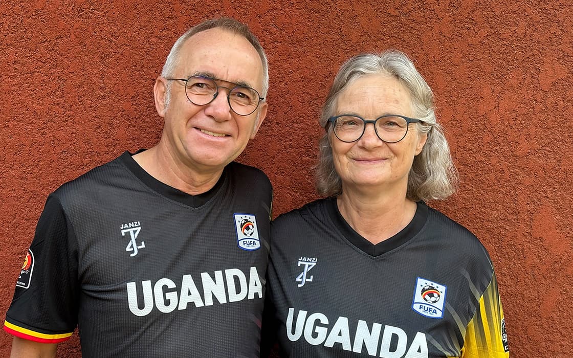 Martin Auch und Tabea Auch, in passenden schwarzen Uganda-Fußballtrikots, stehen lächelnd vor einer strukturierten roten Wand.