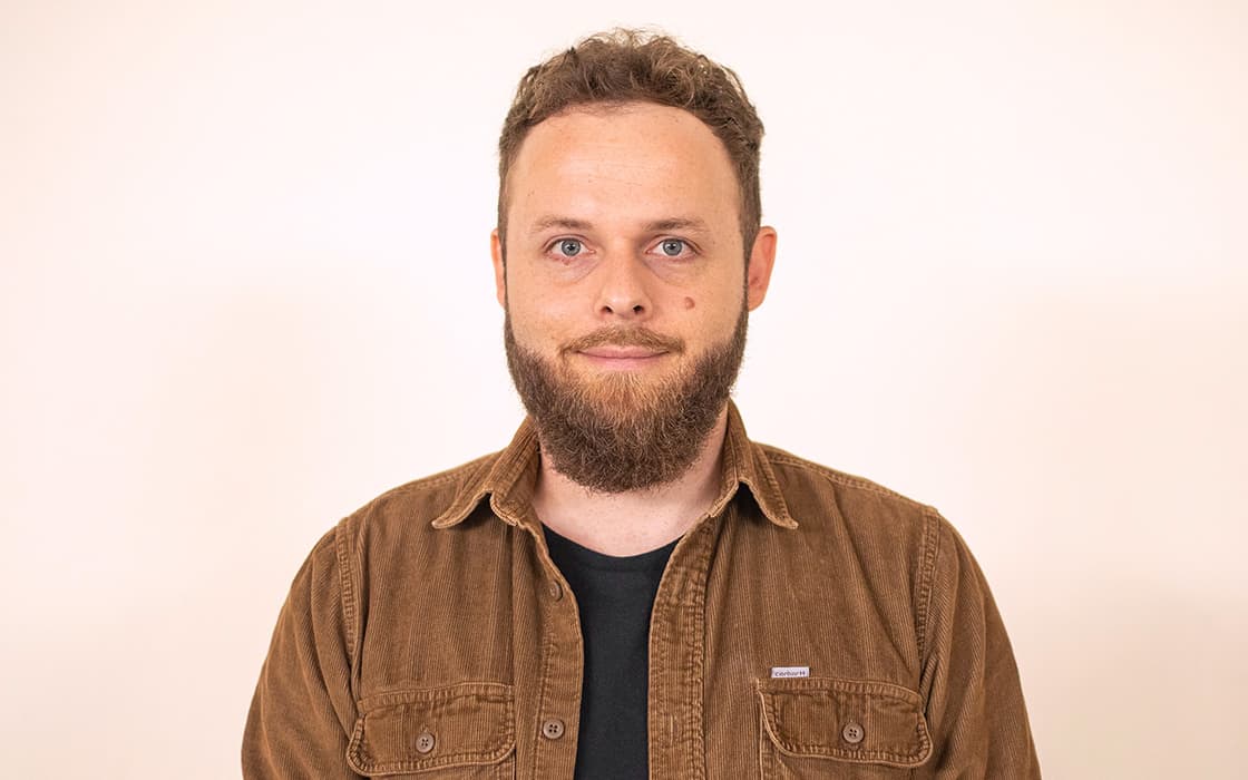 Benjamin Linke, ein Mann mit kurzen lockigen Haaren und einem Bart, trägt ein braunes Hemd mit Knöpfen vor einem schlichten hellen Hintergrund.