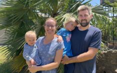 Jana Müller und Markus Müller lächeln mit ihrer vierköpfigen Familie an einem sonnigen Tag im Freien vor einer Palme.