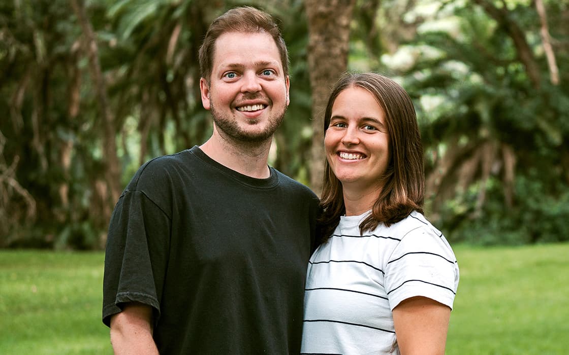 Ein lächelndes Paar, Marc Schwips und Naemi Schwips, steht eng beieinander im Freien mit grünen Bäumen und Gras im Rücken.