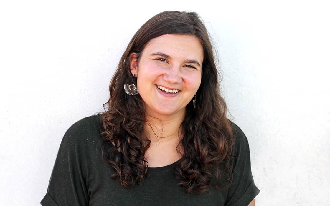 Rebekka Eggeler, mit langen braunen Haaren und einem dunklen Hemd, lächelt vor einem schlichten weißen Hintergrund.