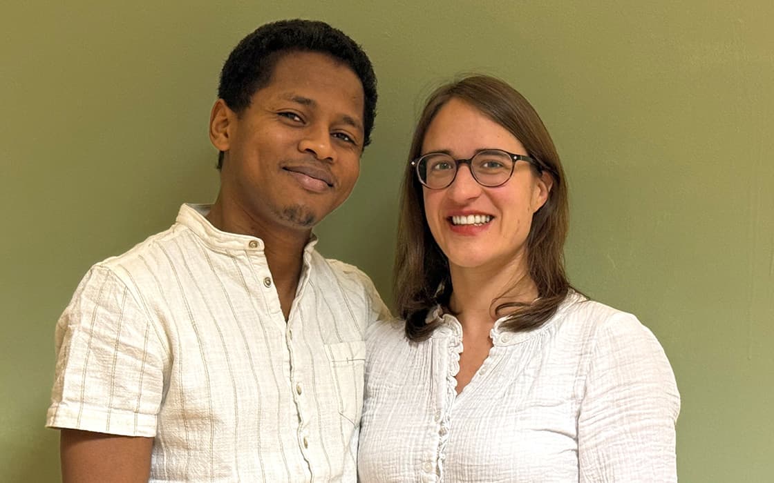 Flavien Fiangoa und Debora lächeln gemeinsam in weißen Hemden vor einem schlichten grünen Hintergrund.