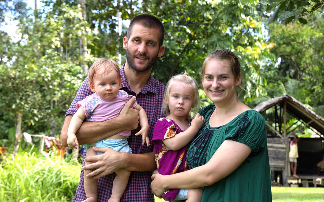Eine lächelnde vierköpfige Familie, darunter Anne Braunmiller, posiert im Freien mit Grünpflanzen und einer Holzhütte im Hintergrund.