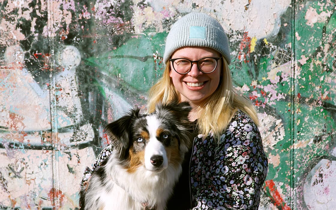 Die lächelnde Maika Hirschfeld mit Brille und Mütze hält einen Australian Shepherd vor einer bunten, mit Graffiti beschmierten Wand.