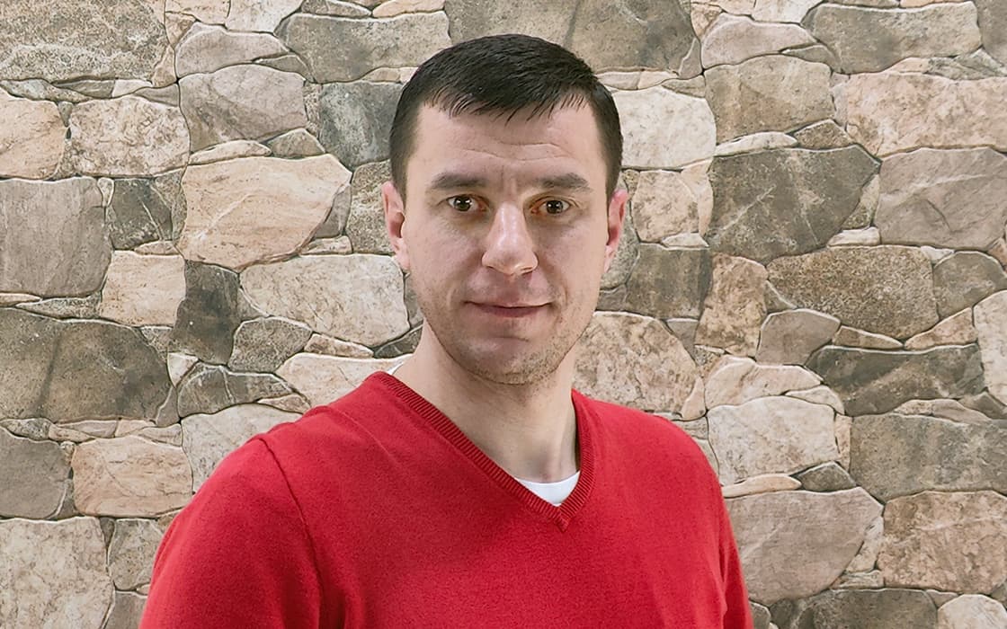 Alexander Schimpf, mit kurzen dunklen Haaren und rotem Pullover, steht vor einer mit Steinen gemusterten Wand und schaut in die Kamera.