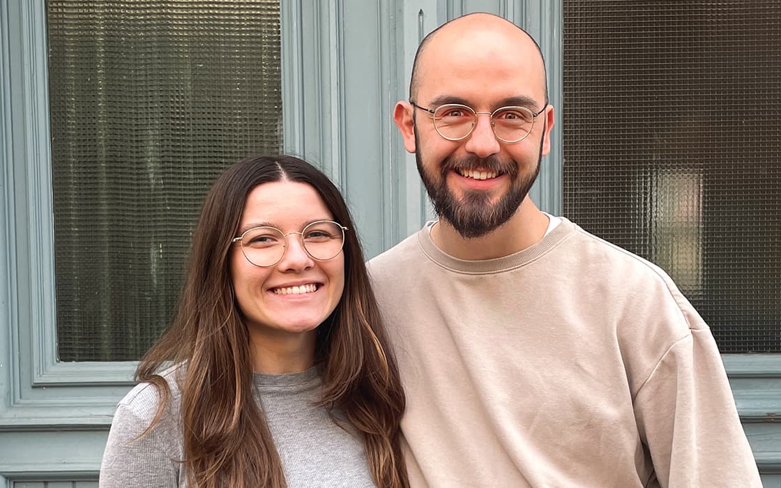 Zwei Personen mit Brillen, die lächelnd dicht beieinander vor hellblauen Holztüren und -fenstern stehen.