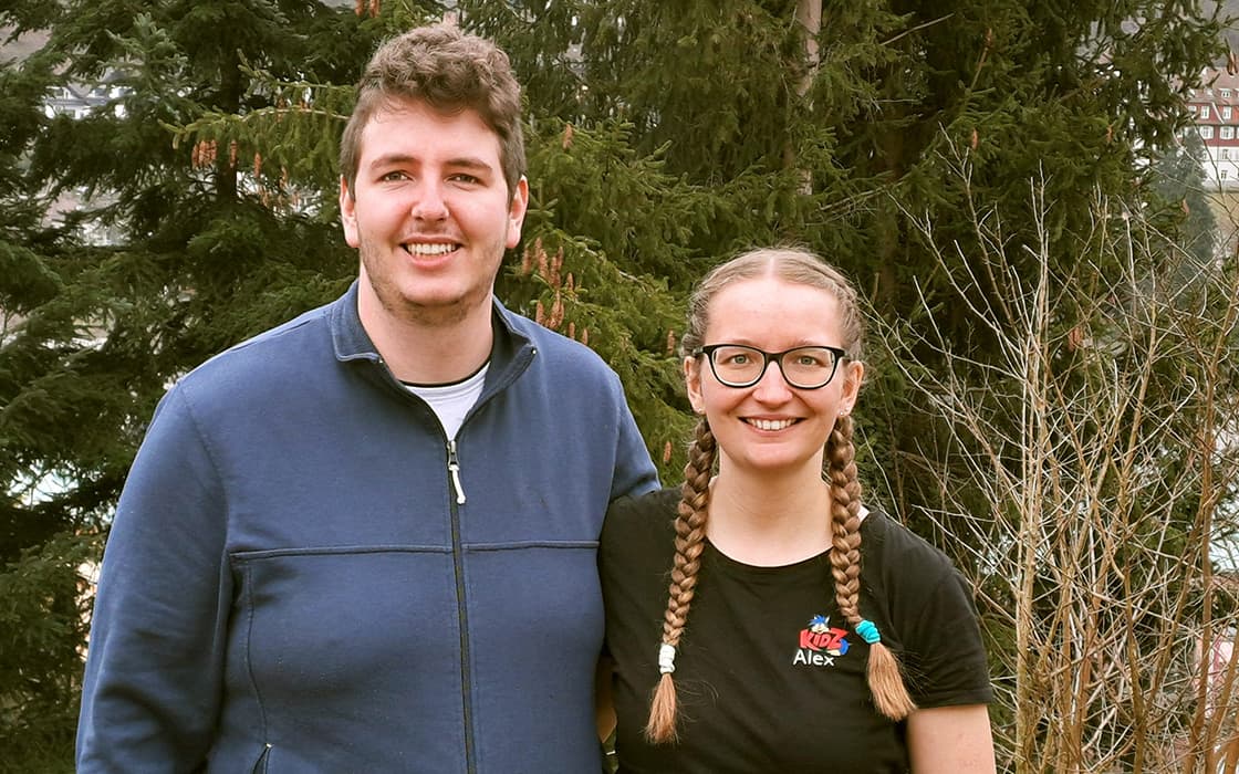 Alexandra und Manuel Veigel lächeln im Freien vor immergrünen Bäumen, die eine mit Brille und Zöpfen, der andere in einer blauen Jacke.