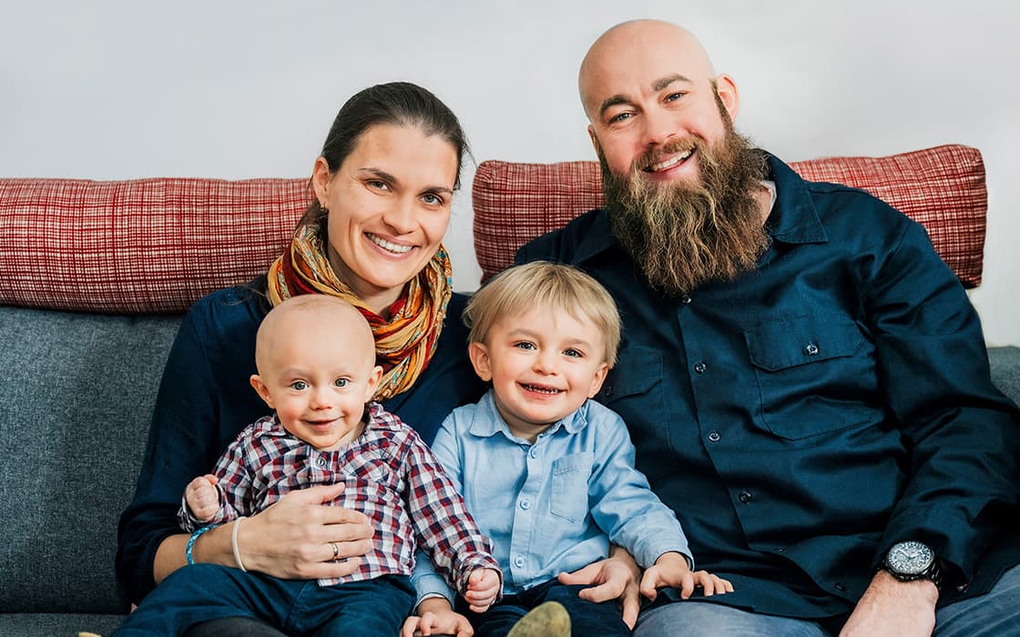 Eine lächelnde vierköpfige Familie sitzt auf einer Couch - Johannes Wagler und Carmen Wagler mit ihren beiden kleinen Kindern.