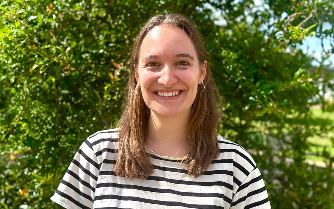 Eine lächelnde Frau, Lena Schilling, mit glattem braunem Haar und gestreiftem Hemd, steht an einem sonnigen Tag im Freien zwischen grünen, belaubten Büschen.