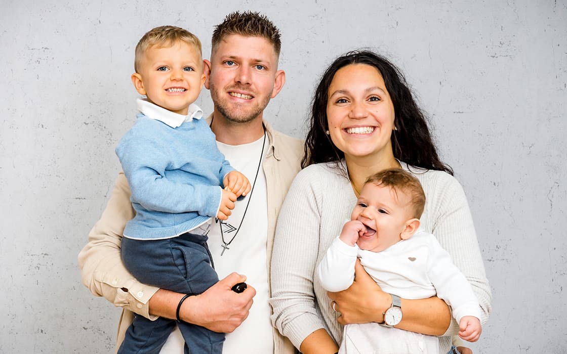Sebastian und Samira Urschitz lächeln, während sie einen kleinen Jungen und ein Baby halten, alle glücklich vor einem hellgrauen Hintergrund.