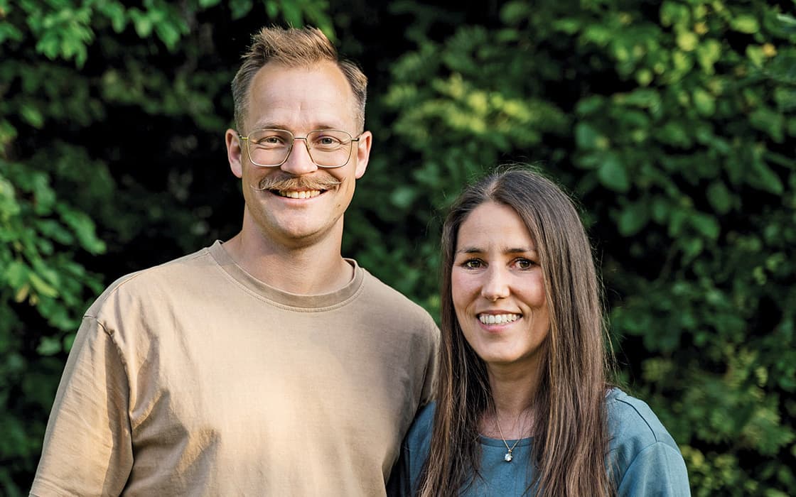 Bernd Stamm und Esther Stamm stehen lächelnd zusammen im Freien vor grünen, belaubten Sträuchern.