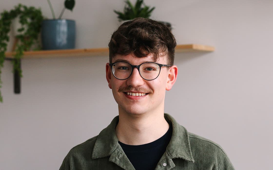 Eine lächelnde junge Person mit Brille und Schnurrbart steht in einem Haus mit Pflanzen auf einem Regal im Hintergrund.