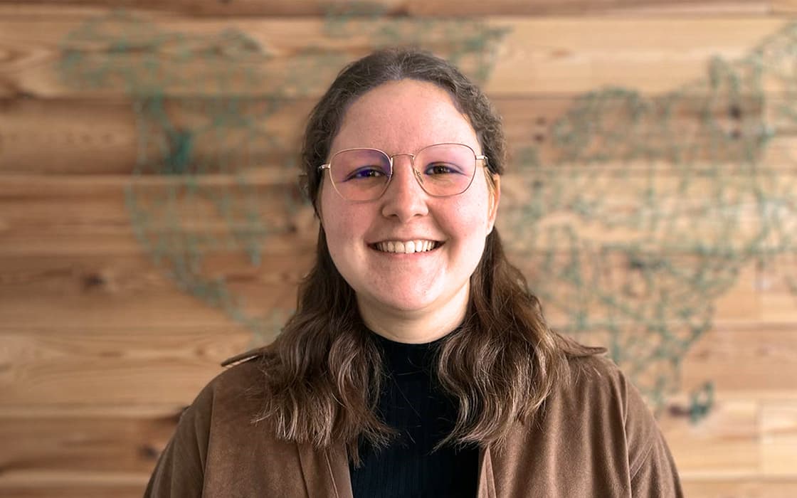 Die lächelnde Paula Otterbach mit Brille steht vor einer Holzwand mit einem Kartenmotiv im Hintergrund.