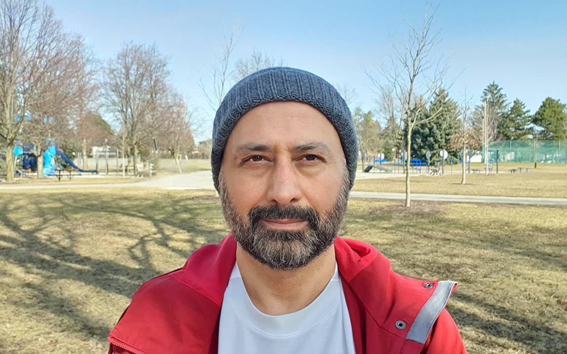 Ein Mann mit grauer Mütze und roter Jacke steht an einem klaren Tag draußen in einem Park.