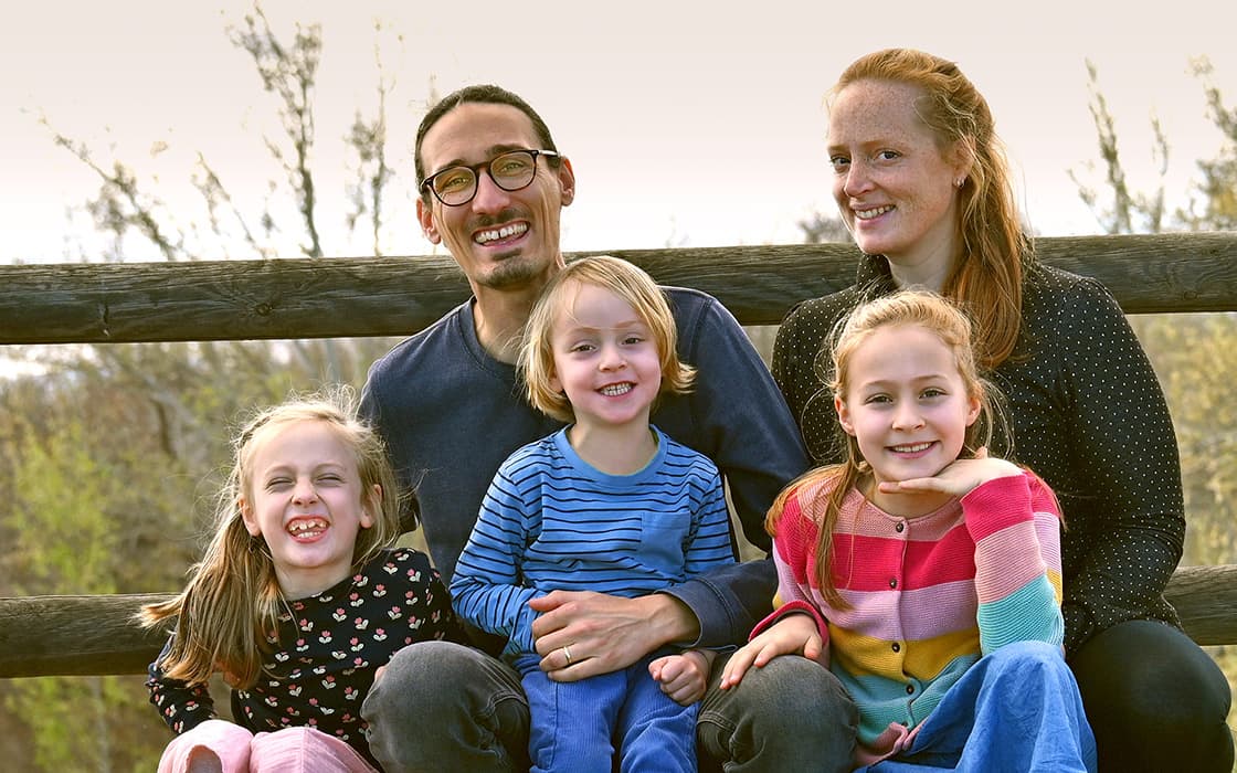 Eine lächelnde fünfköpfige Familie, darunter Mirjam Schmückle und David Schmückle, posiert gemeinsam im Freien an einem Holzzaun.