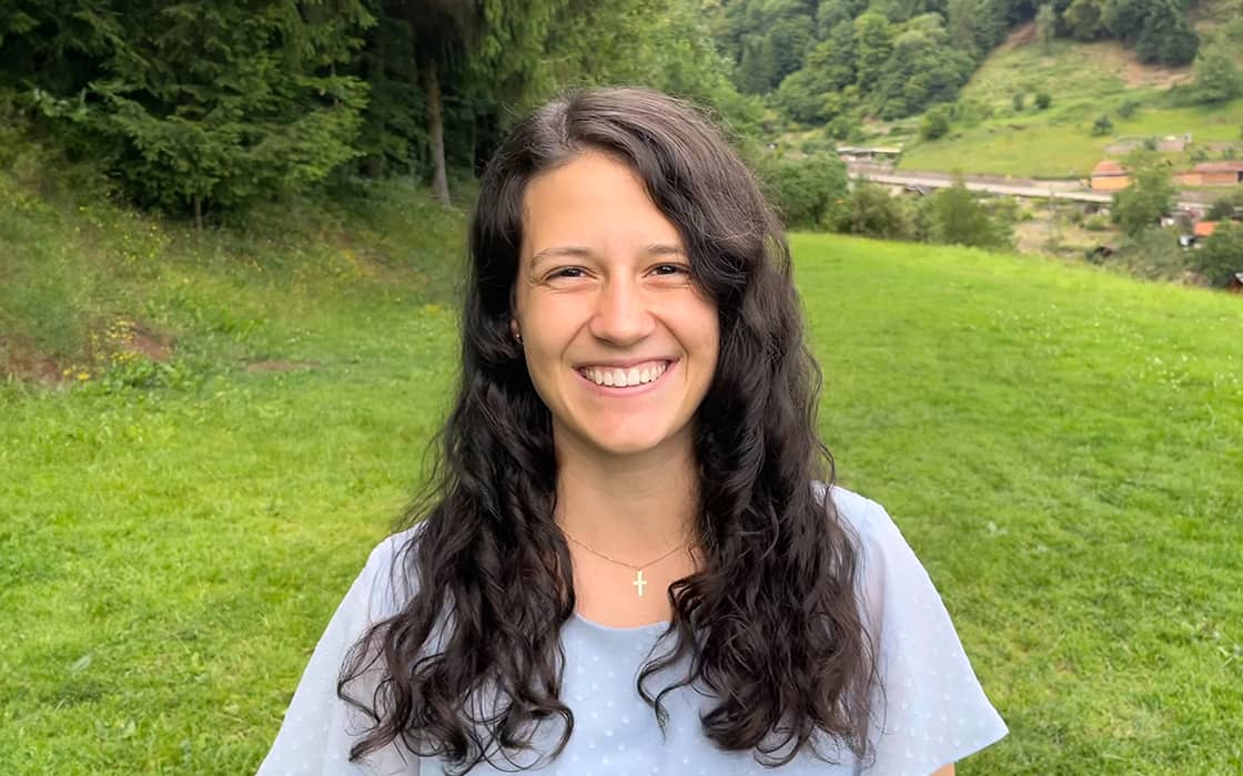 Eva Reichert, eine Frau mit langen dunklen Haaren, lächelt, während sie auf einer Wiese mit Bäumen und Hügeln im Hintergrund steht.