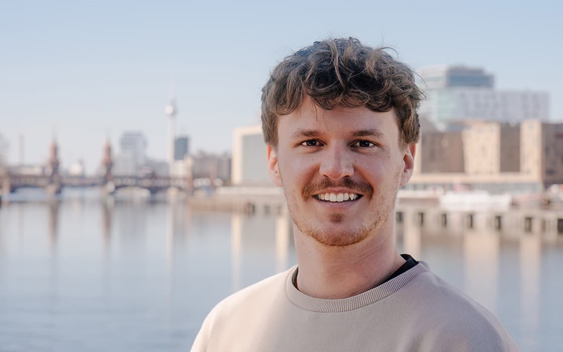 Ein lächelnder Mann mit gewelltem Haar steht an einem klaren Tag an einem Flussufer mit einer Stadtsilhouette im Hintergrund.