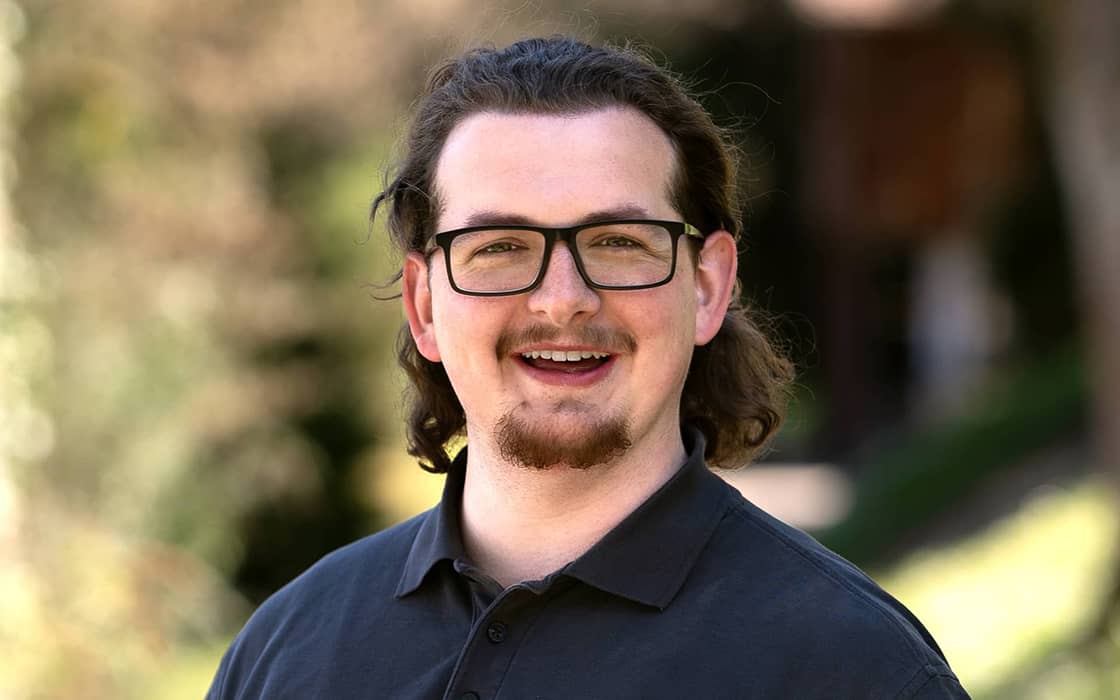 Der lächelnde Benjamin Rüb mit Brille und Ziegenbart, der ein dunkles Hemd trägt, steht im Freien vor einem unscharfen Hintergrund.