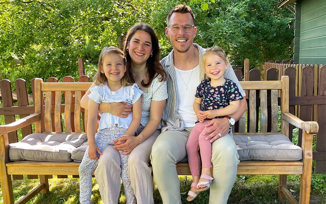 Eine vierköpfige Familie, darunter Seda Battermann und Christian Battermann, lächelt auf einer Holzbank in einem sonnigen Hinterhof.