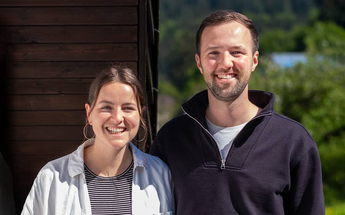 Paul und Anna Frank lächeln im Freien, dicht beieinander stehend, mit Grünzeug und einer Holzwand im Hintergrund.