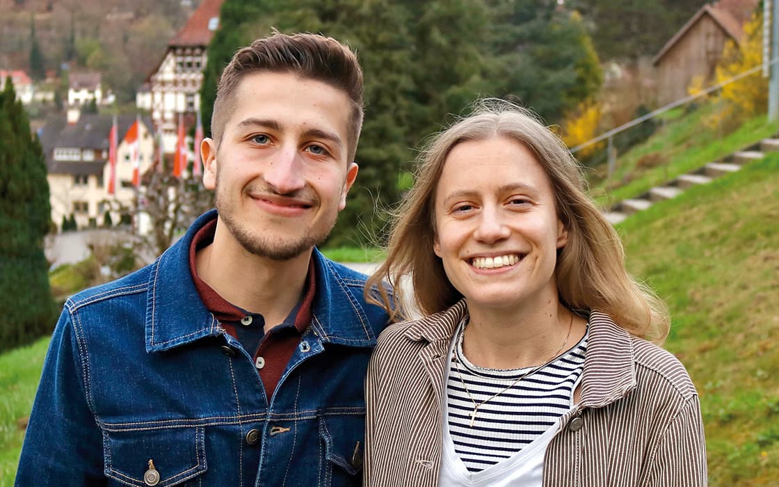 Muriel Boucherit und Elias Boucherit lächeln im Freien, dicht beieinander stehend, mit Grünflächen und Gebäuden im Hintergrund.