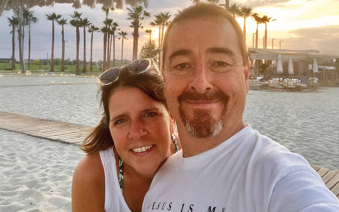 Das lächelnde Paar Christine Abel und Volker Abel macht ein Selfie an einem Sandstrand bei Sonnenuntergang, mit Palmen im Hintergrund.