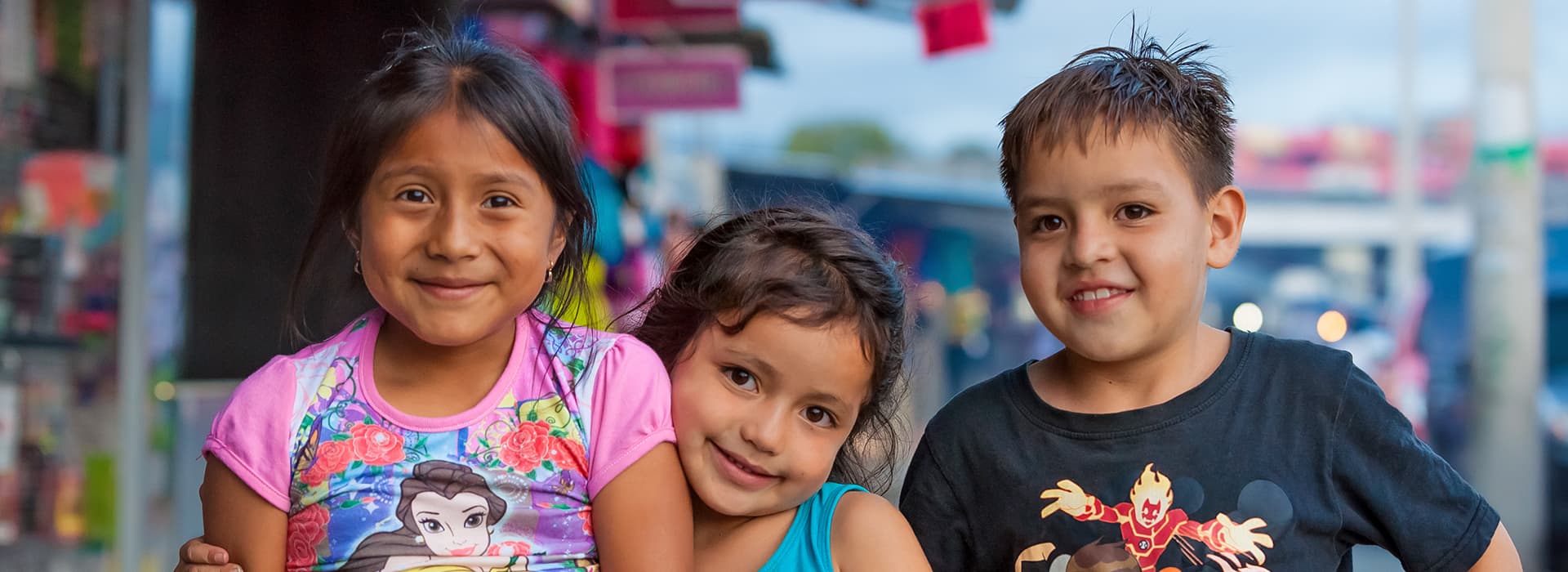 Drei lächelnde Kinder, darunter Kathy Bruce und Kevin Bruce, stehen dicht beieinander mit bunten Schildern hinter ihnen.