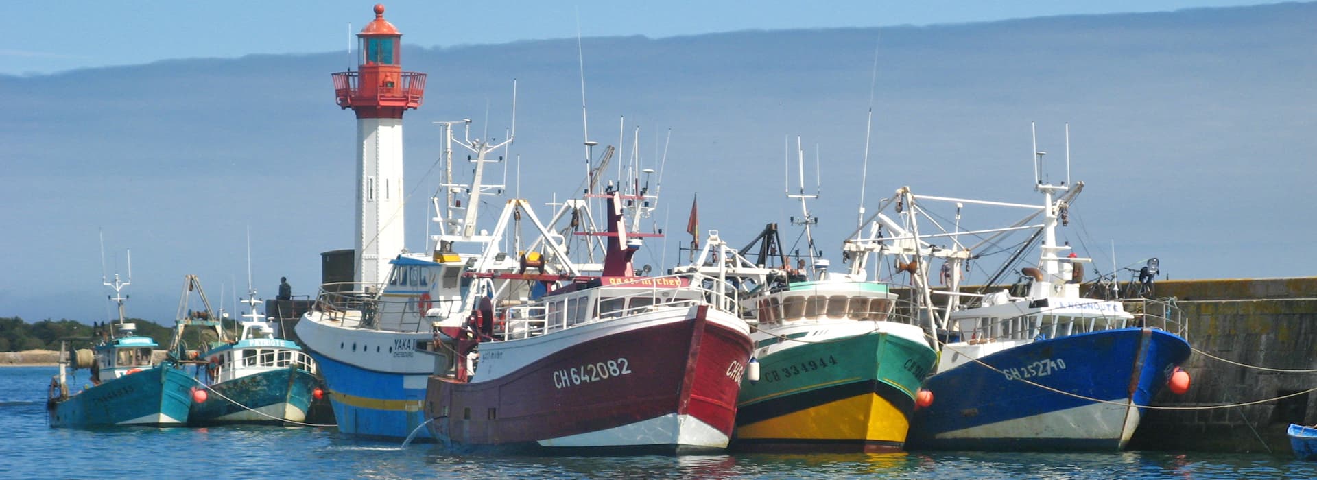 An einem sonnigen Tag liegen bunte Fischerboote in der Nähe eines rot-weißen Leuchtturms, gemalt von Tina Eckstein, vor Anker.