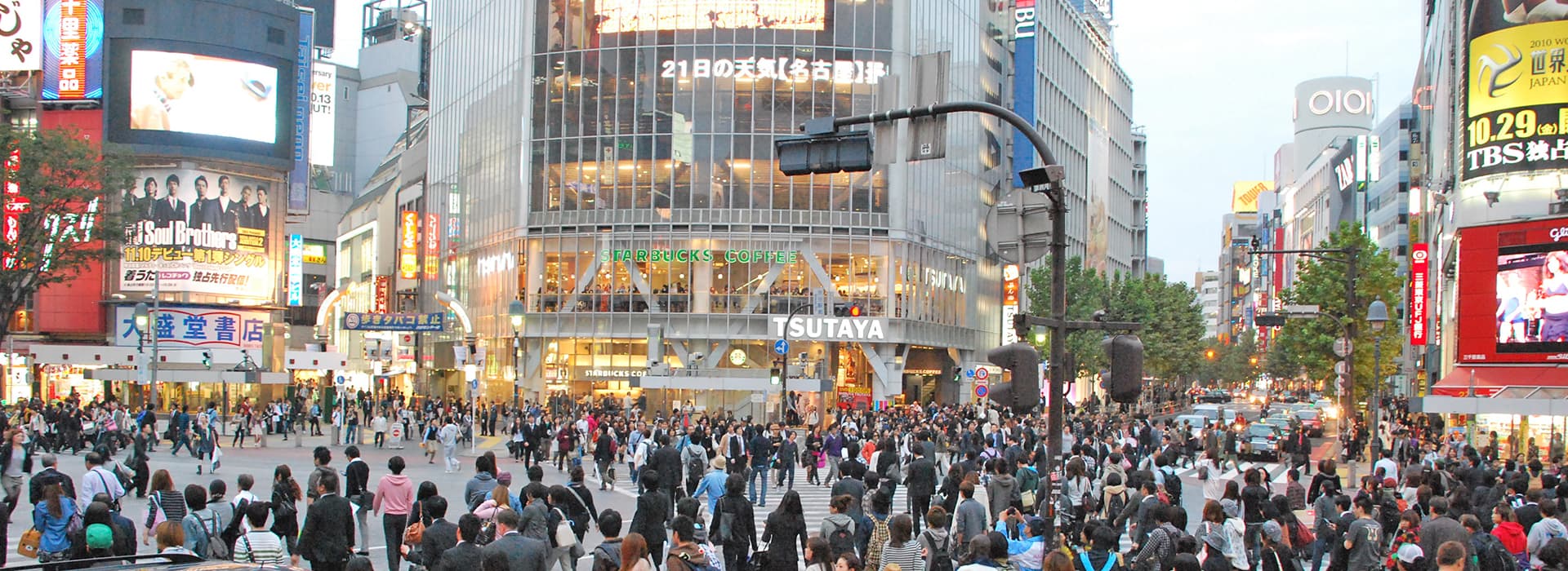 Eine große Menschenmenge überquert eine belebte Kreuzung in einer Stadt, während Thomas Beck die hellen Schilder und hohen Gebäude betrachtet.