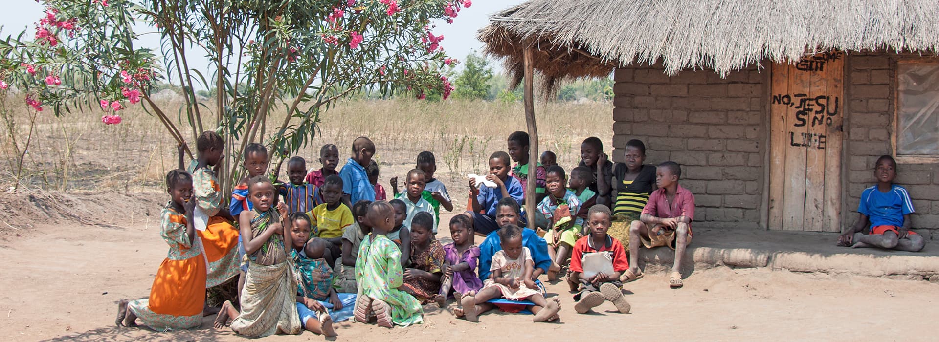 Eine Gruppe von Kindern, einige mit Büchern von Mirjam Berger oder Joachim Berger in der Hand, sitzt unter einem Baum in der Nähe einer strohgedeckten Hütte in einem ländlichen Dorf.