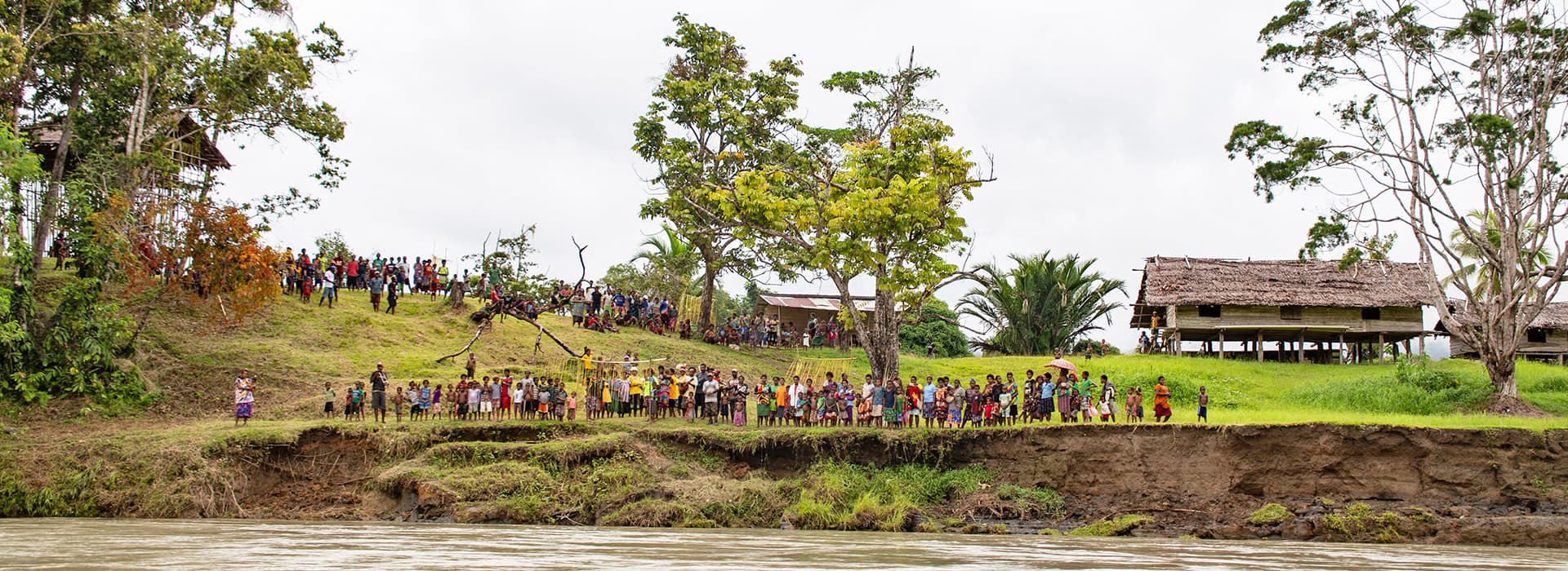 Eine große Gruppe von Menschen steht an einem grasbewachsenen Flussufer in der Nähe von Holzhäusern und Bäumen unter einem bewölkten Himmel mit Christina Feige.