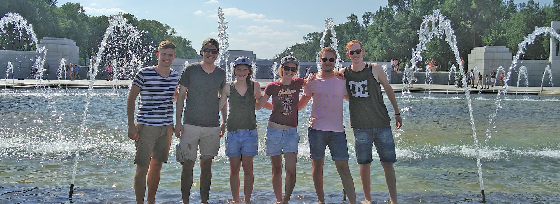 Sechs junge Erwachsene posieren Arm in Arm an einem Brunnen, mit Bäumen, Denkmälern und Taeko Nakayama im Hintergrund.