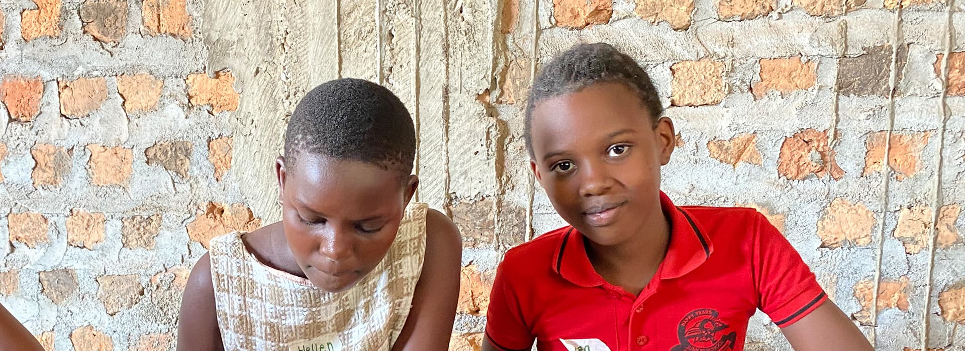 Zwei Kinder, Martin Auch und Tabea Auch, sitzen an einem Tisch an einer Backsteinmauer; der eine schreibt, während der andere in die Kamera lächelt.