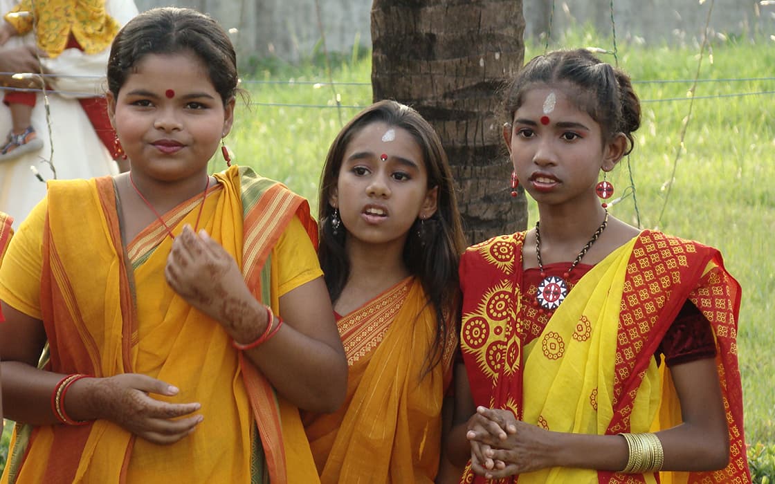 Drei Mädchen in traditioneller gelber und roter indischer Kleidung stehen im Freien, tragen Bindis und Schmuck und schauen nach vorne.